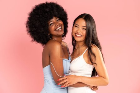 Photo for Fashionable asian woman posing with her friend with afro hairstyle. Cheerful, elegant girls standing together, looking at the camera and smiling happily. Copy space. Real people. Pink pastel studio background. - Royalty Free Image