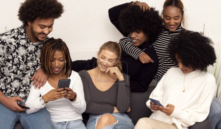 Photo for Group of cheerful young men and women laughing while looking at smartphone and watching funny videos on social media, at home. Multiethnic people sitting on sofa having fun together. - Royalty Free Image