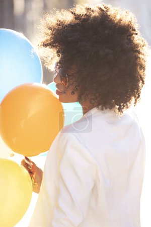 Foto de Mujer afro sonriente disfrutando de un día soleado al aire libre, sosteniendo globos de colores en la mano. Momentos felices. - Imagen libre de derechos