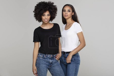 Foto de Dos hermosas chicas afroamericanas posando en el estudio en jeans y camisetas básicas casuales, mirando y sonriendo a la cámara. - Imagen libre de derechos