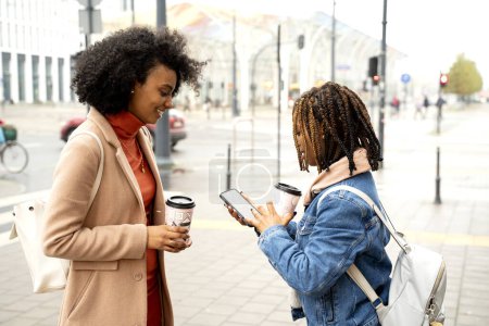 Photo for Two beautiful young african woman friends drinking coffee outdoor, smiling, talking and walking city street. Tourists using phone with map. Real people emotions. - Royalty Free Image