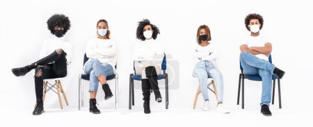 Téléchargez les photos : Groupe multi-ethnique de jeunes portant des masques, assis sur des chaises et regardant la caméra. - en image libre de droit