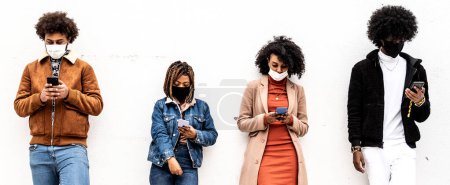 Foto de Grupo africano de amigos que mantienen la distancia al aire libre, de pie sobre la pared con la máscara facial cubierta. Cada persona está mirando el teléfono móvil y viendo nuevo contenido en las redes sociales. - Imagen libre de derechos