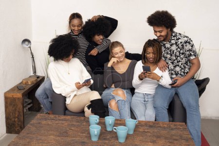 Photo for Group of cheerful young men and women laughing while looking at smartphone and watching funny videos on social media, at home. Multiethnic people sitting on sofa having fun together. - Royalty Free Image