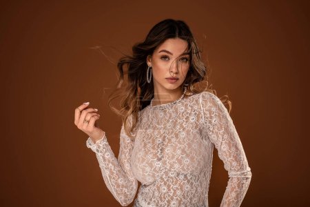 Photo for Beautiful young natural woman with delicate makeup in romantic lace dress and fashionable jewelry posing on studio background. Bride to be with long wavy hair. A lot of copy space on the brown studio background. - Royalty Free Image