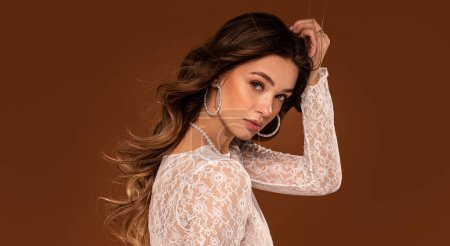 Photo for Beautiful young natural woman with delicate makeup in romantic lace dress and fashionable jewelry posing on studio background. Bride to be with long wavy hair. A lot of copy space on the brown studio background. - Royalty Free Image