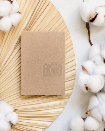 Photo for Blank kraft paper card on dried palm leaf with cotton flowers top view, mockup. Romantic scene with vertical card. Bohemian pastel greeting or wedding Invitation - Royalty Free Image