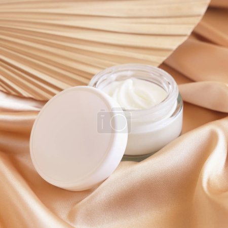 Photo for Opened cosmetic jar with white lid on beige satin fabric and dried palm leaf close up, mockup. Boho scene with glass cream jar. Bohemian or close to nature scen - Royalty Free Image