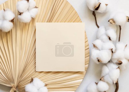 Foto de Blank card on dried palm leaf with cotton flowers top view, mockup. Romantic scene with square paper card. Bohemian pastel greeting or wedding Invitation - Imagen libre de derechos