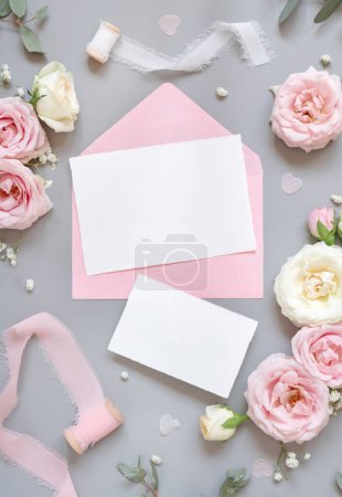 Téléchargez les photos : Paper cards between light pink roses and silk ribbons on grey top view,  wedding mockup. Romantic scene with blank vertical card and pastel flowers flat lay.. Valentines, Spring or Mothers day concept - en image libre de droit