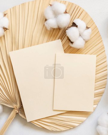 Foto de Tarjetas en blanco en hoja de palma seca con flores de algodón vista superior, maqueta. Escena romántica con tarjetas de papel verticales. Bohemian pastel saludo o boda Invitación - Imagen libre de derechos