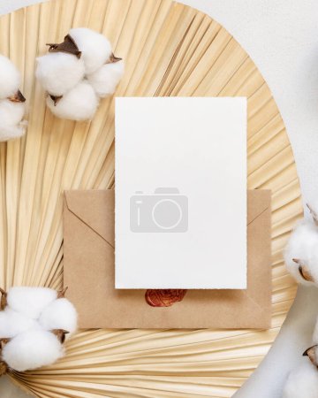 Téléchargez les photos : Blank card and envelope on dried palm leaf with cotton flowers top view, mockup. Romantic scene with vertical paper card. Bohemian pastel greeting or wedding Invitation - en image libre de droit