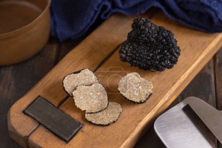 Photo for Whole and sliced black truffles mushroom on wooden board on dark brown table near blue napkin, close up. Culinary delight ingredient. - Royalty Free Image