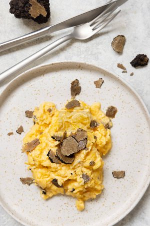 Téléchargez les photos : Scrambled eggs with sliced fresh black truffles from Italy served in a plate near fork and knife top view. Italian cousine, gourmet breakfast - en image libre de droit