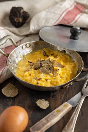 Foto de Scrambled eggs with sliced fresh black truffles from Italy served in a a frying pan near fork and knife on wood close up. Italian cousine, gourmet breakfast - Imagen libre de derechos