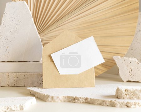 Photo for Blank card and envelope near beige travertine stones against dry beige palm leaf close up, mockup. Boho scene with horizontal paper card. Bohemian greeting or wedding Invitation - Royalty Free Image