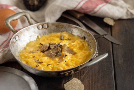 Photo for Scrambled eggs with sliced fresh black truffles from Italy served in a a frying pan near fork and knife on wood close up. Italian cousine, gourmet breakfast - Royalty Free Image