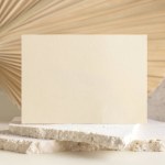 Beige Blank card near beige travertine stones against dry beige palm leaf close up, mockup. Boho scene with horizontal paper card. Bohemian or close to nature greeting or wedding Invitation. Minimal compositio