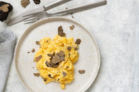 Foto de Scrambled eggs with sliced fresh black truffles from Italy served in a plate near fork and knife top view. Italian cousine, gourmet breakfast - Imagen libre de derechos