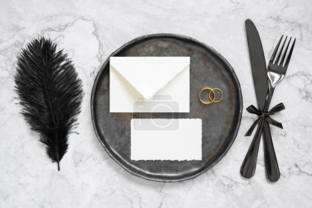 Photo for Black wedding table setting with a blank envelope, card and two rings top view, mockup. Elegant flat lay with black feather and cutlery, wedding or holiday stationery template - Royalty Free Image