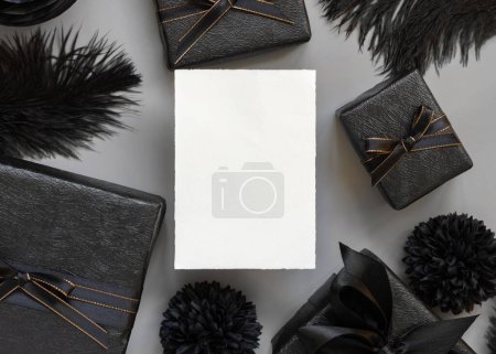 Photo for Blank card between Wrapped Gift Boxes, black paper flowers and feathers, mockup, copy space. Dark composition with vertical paper card for Birthday, Anniversary or black party present - Royalty Free Image