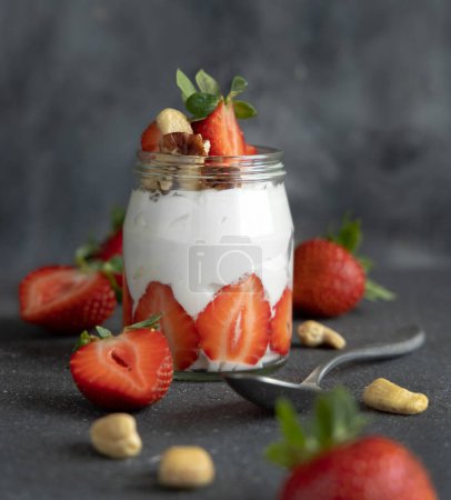 Photo for Greek yogurt, nuts and strawberries in a glass jar on grey concrete table with a spoon, close up. Healthy breakfast with fruits and nuts. Great way to eat protein - Royalty Free Image