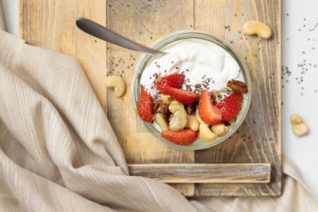 Photo for Greek yogurt, nuts and strawberries in a glass jar on a wooden tray with napkinn, top view. Healthy breakfast with fruits and nuts. Great way to eat protein - Royalty Free Image