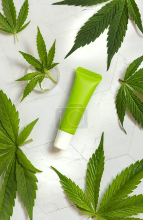 Green blank Cream tube near cannabis sativa leaves on a marble table top view. Cosmetic Mockup, Copy space. Organic skincare beauty product. Eco friendly body or hand cream with hem