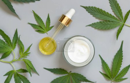 Photo for Pipette with CBD oil and opened cream jar near green cannabis leaves on grey table top view. Organic skincare beauty products, natural cosmetic - Royalty Free Image