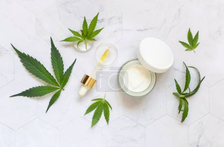 Photo for Pipette with CBD oil and opened cream jar with white lid near green cannabis leaves on white marble table top view. Organic skincare beauty products, natural cosmetic - Royalty Free Image