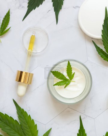 Photo for Pipette with CBD oil and opened cream jar with white lid near green cannabis leaves on white marble table top view. Organic skincare beauty products, natural cosmetic - Royalty Free Image