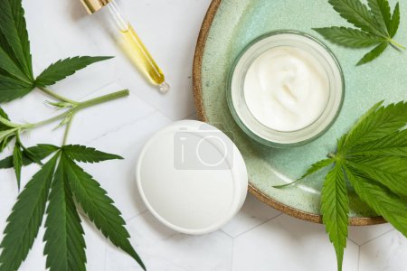 Photo for Pipette with oil and opened cream jar with white lid  near green cannabis leaves on plate on white marble table top view, mockup. Organic skincare beauty products, natural cosmetic - Royalty Free Image