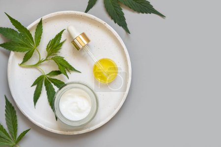 Cream jar and pipette near green cannabis sativa leaves on plate on grey table top view, copy space. Organic skincare beauty product. Eco friendly CBD Cosmetic