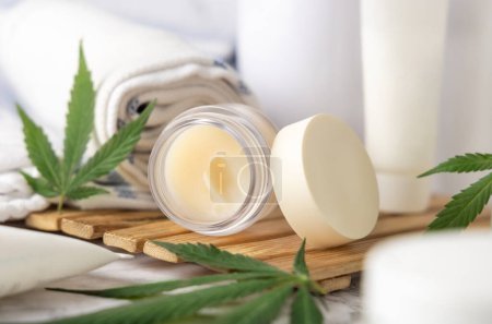 Photo for Jar with CBD lip balm and green cannabis leaves near bottles and towel in bathroom close up on a wooden tray. Organic skincare product. Alternative cosmetic - Royalty Free Image