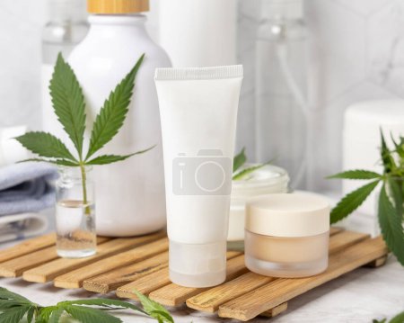 Téléchargez les photos : Tube blanc blanc crème près de feuilles de cannabis vert sur plateau en bois dans la salle de bain près d'autres bouteilles, gros plan, maquette. Produits de beauté bio pour la peau, cosmétiques naturels CBD - en image libre de droit