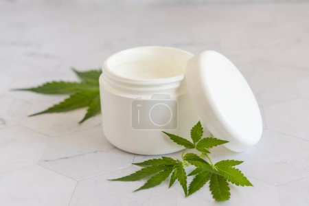 Photo for Opened white cream jar with a lid near green cannabis sativa leaves on a marble table. Cosmetic Mockup, Copy space. Organic skincare beauty product. Eco friendly body or hand cream with hem - Royalty Free Image