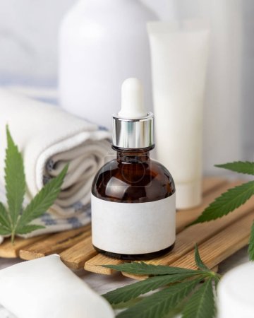 Photo for Dropper bottle with blank label and green cannabis leaves near bottles and towel close up in bathroom, mockup. Organic CBD cosmetic. - Royalty Free Image