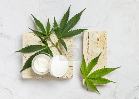 Foto de Frasco de crema abierto con tapa en blanco y hojas de cannabis verde en vista superior de piedra beige. Cosmética ecológica ecológica CBD. Estilo de vida alternativo - Imagen libre de derechos