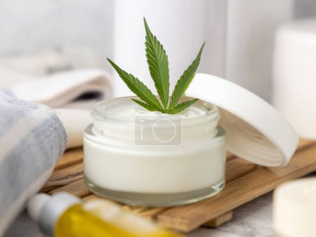 Téléchargez les photos : Pot de crème ouvert avec des feuilles de cannabis vert sur le plateau fermer dans la salle de bain. Produits de beauté bio pour la peau, cosmétiques alternatifs CBD - en image libre de droit