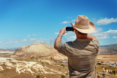 Photo for Man with shirt and cowboy hat taking photos with mobile phone during trekking in a sunny day, back view. Man took a stop during walking in mountains to see beautiful view - Royalty Free Image