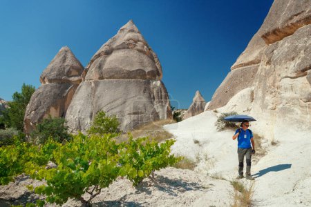 Téléchargez les photos : Homme avec parapluie marchant entre des formations géologiques uniques en Cappadoce contre le ciel bleu en été. Zone touristique populaire dans Goreme, Nevsehir, Turke - en image libre de droit