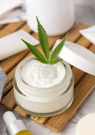 Téléchargez les photos : Pot de crème ouvert avec des feuilles de cannabis vert sur le plateau fermer dans la salle de bain. Produits de beauté bio pour la peau, cosmétiques alternatifs CBD - en image libre de droit