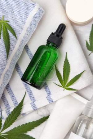 Photo for Green glass dropper bottle near green cannabis leaves on towel top view, mockup. Organic skincare beauty products, alternative CBD cosmetic - Royalty Free Image