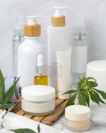 Téléchargez les photos : Pots, tubes et bouteilles cosmétiques vierges près des feuilles de cannabis vert sur un plateau en bois dans la salle de bain près d'autres bouteilles, gros plan, maquettes. Produits de beauté bio pour la peau, cosmétiques naturels CBD - en image libre de droit