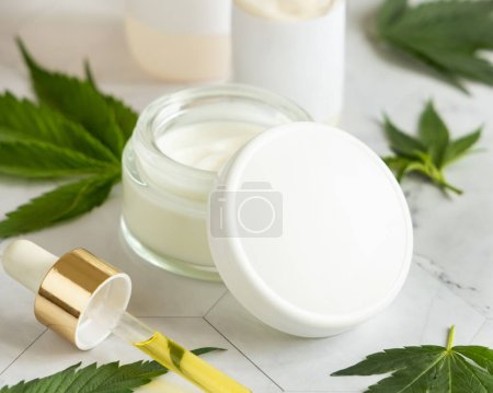 Photo for Cream jar with a blank lid and pipette with CBD oil near cannabis leaves on a marble table. Cosmetic Mockup, Copy space. Organic skincare beauty product. Eco friendly body or hand cream with hem - Royalty Free Image