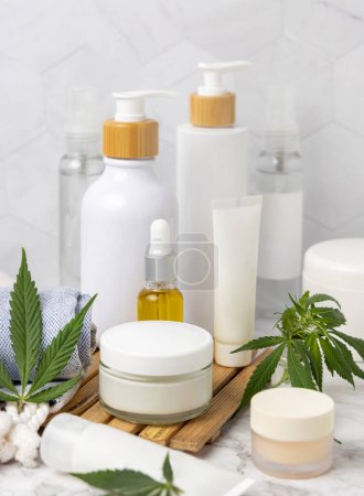 Foto de Frascos cosméticos en blanco, tubos y botellas cerca de hojas de cannabis verde en bandeja de madera en el baño cerca de otras botellas, de cerca, maqueta. Productos orgánicos de belleza para el cuidado de la piel, cosméticos naturales de CBD - Imagen libre de derechos