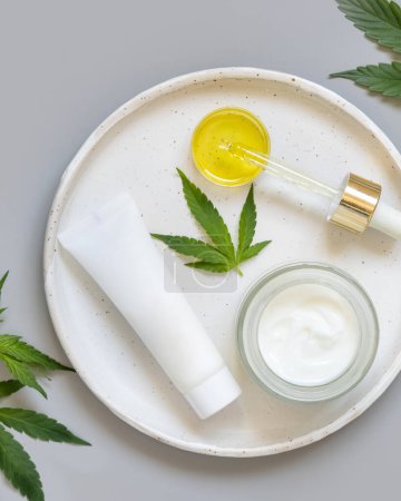 Photo for Blank cosmetic tube, jar and pipette near green cannabis sativa leaves on plate on a grey table top view. Mockup, Copy space. CBD Organic skincare beauty products. - Royalty Free Image