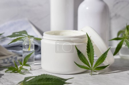 Photo for Opened white cream jar near green cannabis leaves in bathroom near other bottles, close up, mockup. Organic skincare beauty products, natural CBD cosmetic - Royalty Free Image