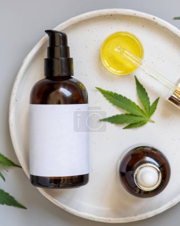 Photo for Blank cosmetic bottles with blank label and pipette with oil near green cannabis sativa leaves on plate on a grey table, top view, Mockup, Copy space. CBD Organic skincare beauty products. - Royalty Free Image