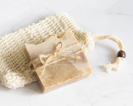 Foto de Barra de jabón beige hecha a mano en bolsa de ahorro de jabón de sisal en la mesa de mármol blanco de cerca, espacio de copia. Productos herbarios naturales para el cuidado facial y corporal - Imagen libre de derechos